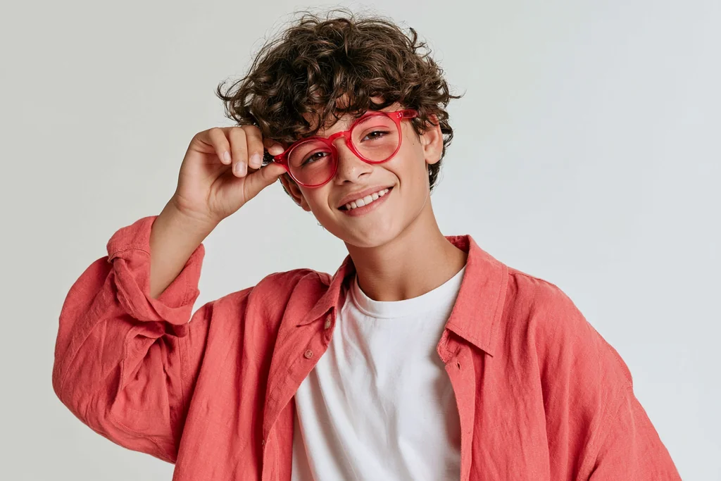 Teenage boy with curly hair in red glasses and an open coral linen shirt, light gray studio