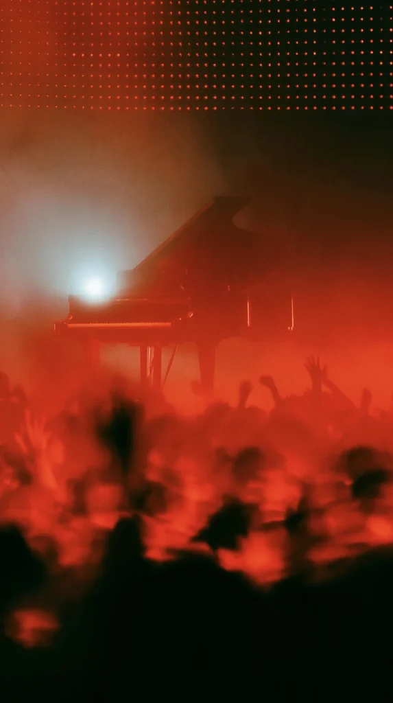 A grand piano silhouetted in strobe and haze over a red-lit club crowd