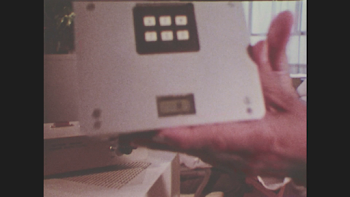 1970s: Hands move object and place it on equipment. Two men look ...