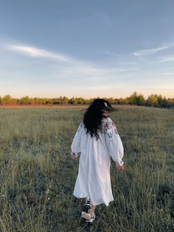 A Back View of a Woman in White Dress Walking on Green Grass Field