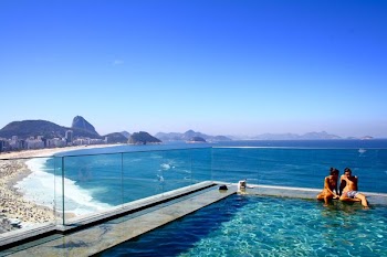 A couple sitting on the edge of a swimming pool overlooking a crowded beach and a deep blue sea
