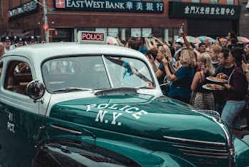 A Crowd Of People Gathered Around A Green Vintage Police Car In The City Street
