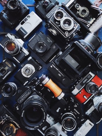 A layflat collection of retro and antique cameras on a blue background like Pentax 6x7, Mamiya RZ67, Holga, Contax, Rolleiflex and Nikon. 