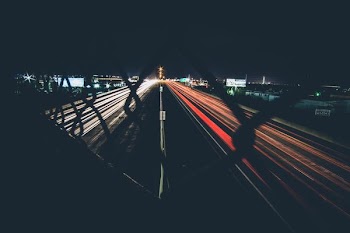 A night-time shot of car trails on a dual highway from behind a fence
