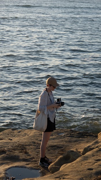 A photograph of a woman holding a camera standing on a  seashore