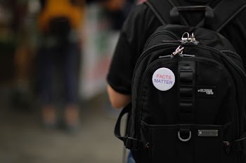 A white“Facts matter” pin on a black backpack on a person's back