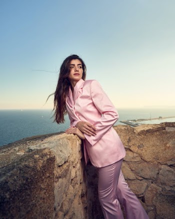 a woman in a pink suit leaning against a stone wall near the ocean