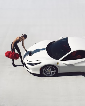 a woman standing next to a white sports car with a red bag on it's hood and her hand on the hood of the car