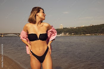 A young woman in a black swimsuit with a pink shirt stands near the water in the city. Portrait of a woman. Brown tint. Relax summer