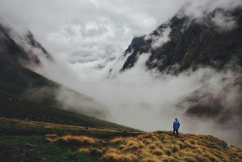 aerial photography of hooded man standing at mountain cliff facing cloudy mountains
