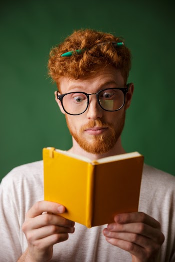 Amazed young red bearded student in glasses reading the notebook with pen in his hair Free Photo