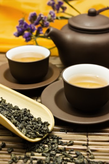Asian tea set with dried green tea on a bamboo surface with a purple flower in the background.