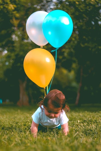 Baby Wearing White T-shirt Holding Three Yellow, Blue, and White Balloons on Green Grass Near Woods