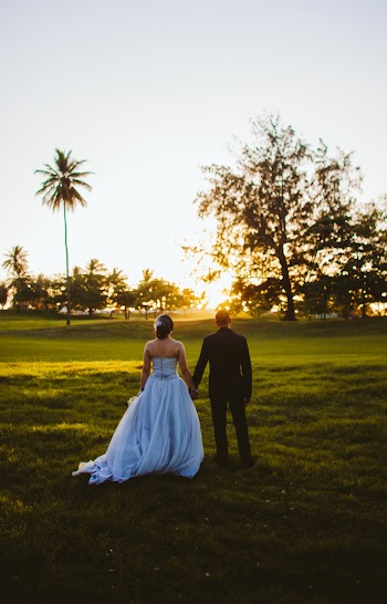 Back View Photo of Bride and Groom Holding Hands While Standing on Grass Field