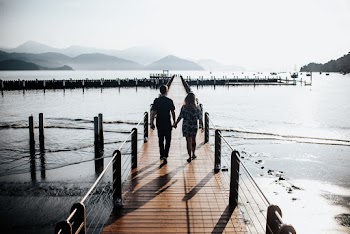 Back View Photo of Couple Holding Hands While Walking Down Wooden Dock