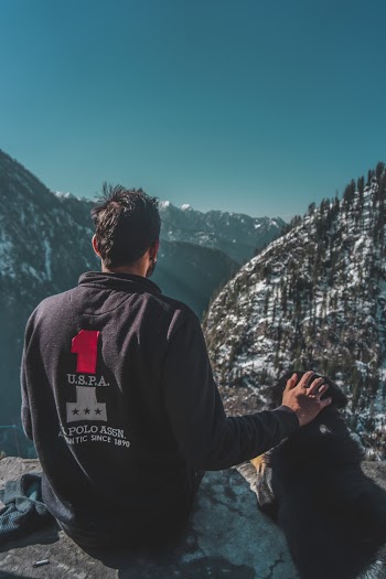 Back View Photo of Man Sitting on Edge of Cliff With His Dog Overlooking Mountains