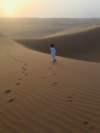 Back View Photo of Muslim Boy in White Thobe Walking Alone on Desert Sand During Golden Hour
