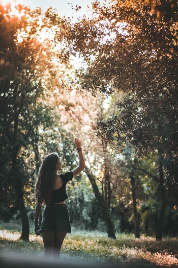 Back View Photo of Smiling Woman in Black Crop Top and Denim Skirt Standing Near Forest Tree With Her Hand Up