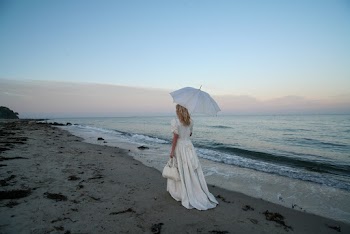 Back View Photo of Woman in White Dress Holding Umbrella and Carrying a Handbag While Walking Alone by the Beach