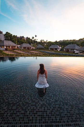 Back View Photo of Woman in White Dress Standing in the Middle of an Infinity Pool Looking at Resort Cottages