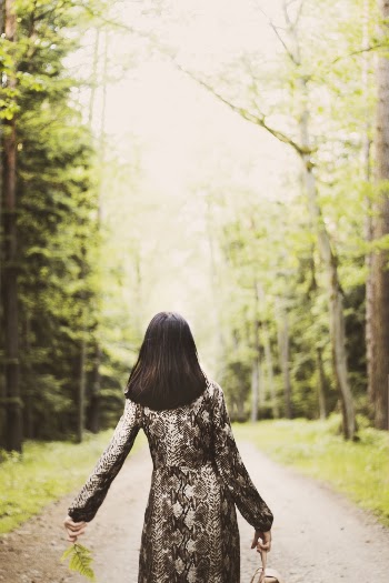Back View Photo of Woman Walking Along Dirt Road Between Trees Carrying Bag
