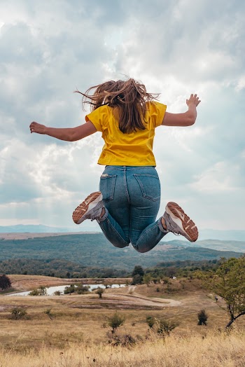 Back view Photo of Woman Wearing Yellow T-shirt and Jeans Jumping on Grass Field