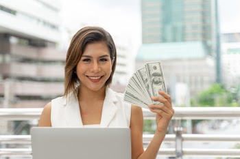 Beautiful girl smiling in business woman clothes using laptop computer and show money us dollar bills in hand Free Photo