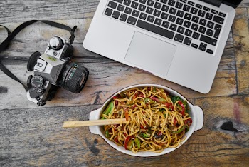 Black and Gray Dslr Camera next to a Laptop and Bowl of Noodles