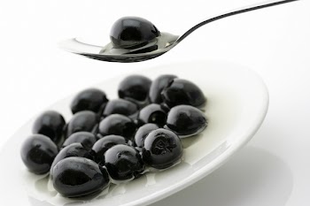 Black olive in spoon over plate with heap of black olives in oil.