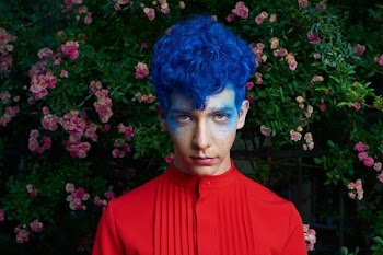 blue haired man standing in front of pink flower plants