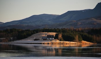 Calm Large Body of Water Across White and Brown House Beside Woods Across Hill