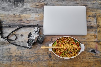 Camera Beside Laptop And Stir Fry Noodles In Bowl On Table