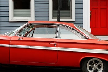 classic red coupe parks beside gray and white wooden house