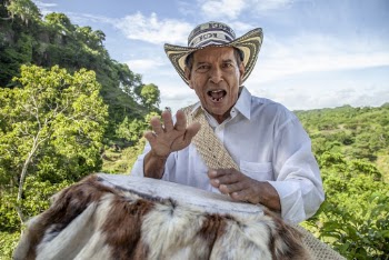 Close-up Of An Elderly Man Wearing A Hat Playing A Percussion Instrument Trimmed With Animal Fur Outdoors