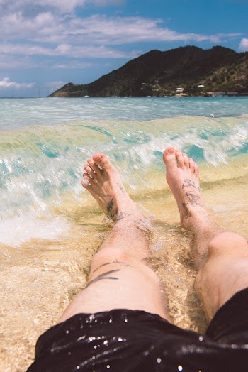 Close-up Of Tattooed Legs And Feet Of A Person Lazing By The Seashore With The Waves Splashing Into Shore