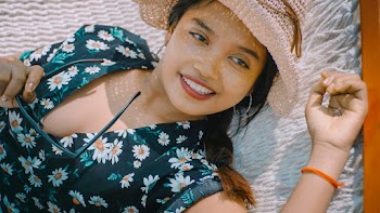 Close-up Photo of Smiling Woman in Black Floral Top and Sun Hat Holding Sunglasses Lying on White Hammock Looking Away