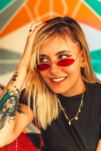 Close-up Photo of Tattooed Woman in Sunglasses Posing with Her Hand Resting on Her Knee and Head