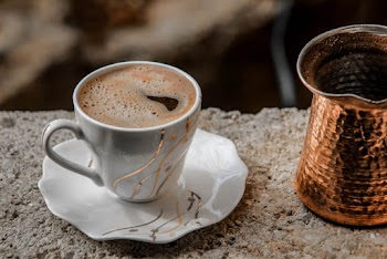 Close-up Photo of White Ceramic Cup and Saucer Set With Coffee Inside Next to a Brass-colored Coffee Pot