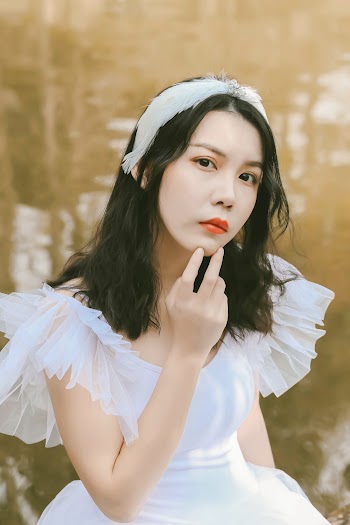 Close-up Photo of Woman in White Dress and White Feather Headband Posing While Sitting Next to Body of Water