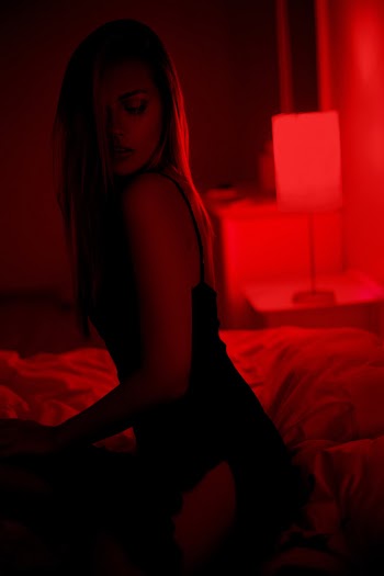 Dark photo of sexy beautiful brunette woman sitting on bed in black dress posing in red interior Free Photo