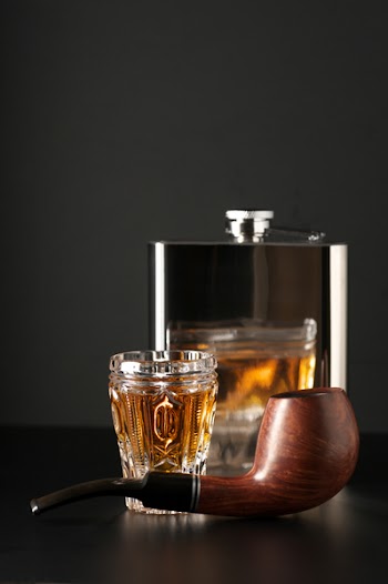Facetted glass of cognac, stainless flask and tobacco pipe on black background.