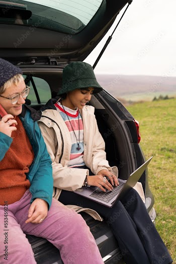 Female hikers sitting in car trunk and using phone and laptop