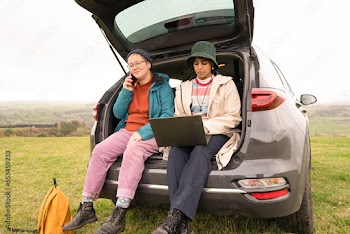 Female hikers sitting in car trunk and using phone and laptop