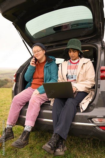 Female hikers sitting in car trunk and using phone and laptop