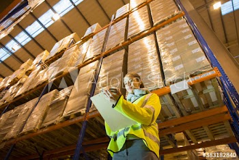 Female worker with clipboard below boxes in distribution warehouse