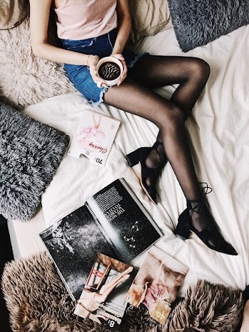 Flatlay Photography of a Woman Holding White Mug With Black Liquid While Lying on a Bed Surrounded by Fur Pillows and Magazines