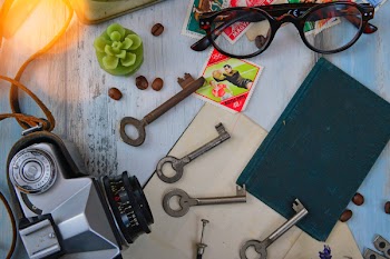 Four Gray Skeleton Keys Next to Gray Camera, Black Book, and Brown Framed Eyeglasses