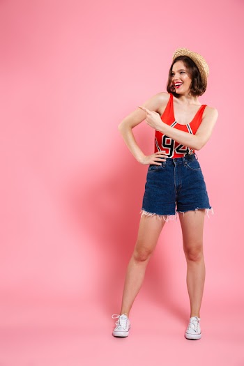 Full length portrait of smiling teen girl in red bodysuit and jeans short standing with hand on her waist, pointing with finger, looking aside Free Photo