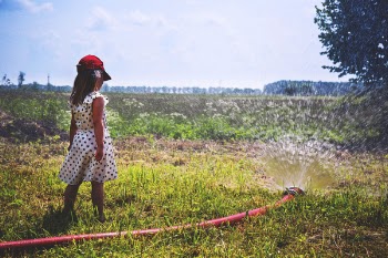 Girl on Green Grass Near Red Hose While Pumping Water during Daytime