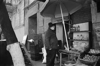 Gray Scale Photo of Man Standing Beside Street With Fruits